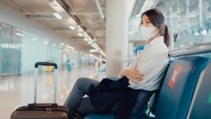 Mulher esperando voo em aeroporto. Ela usa máscara sobre nariz e rosto e ao seu lado há sinalização de distanciamento entre assentos.