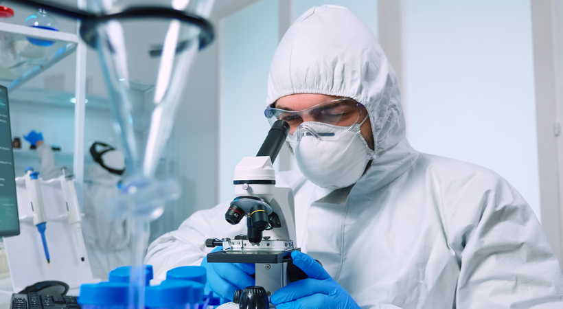 Cientista usando roupa de proteção e luvas em um laboratório enquanto analisa amostras no microscópio.