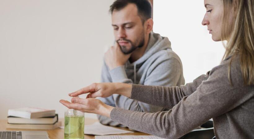 Foto de casal em mesa estudando em livros e cadernos. A mulher está passando álcool em gel enquanto o homem se mantém concentrado. 