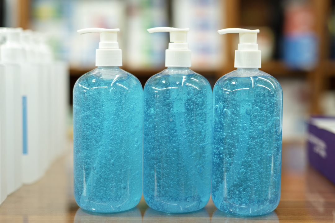 Foto de três frascos de álcool gel em cima de balcão. Ambos são iguais, com pote transparente e conteúdo azul.