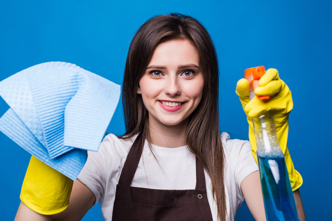 Foto de mulher contente com luvas de limpeza nas mãos. Uma delas segura um pano e a outra um líquido em borrifador.