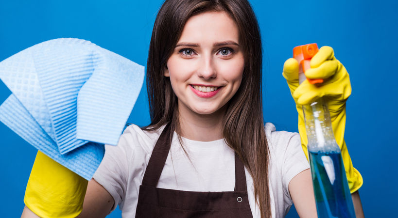 Foto de mulher contente com luvas de limpeza nas mãos. Uma delas segura um pano e a outra um líquido em borrifador. 