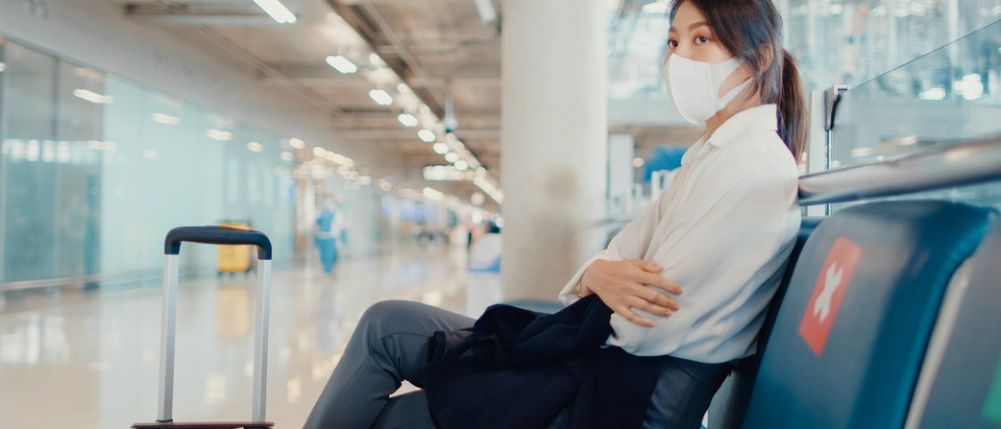 Mulher esperando voo em aeroporto. Ela usa máscara sobre nariz e rosto e ao seu lado há sinalização de distanciamento entre assentos.