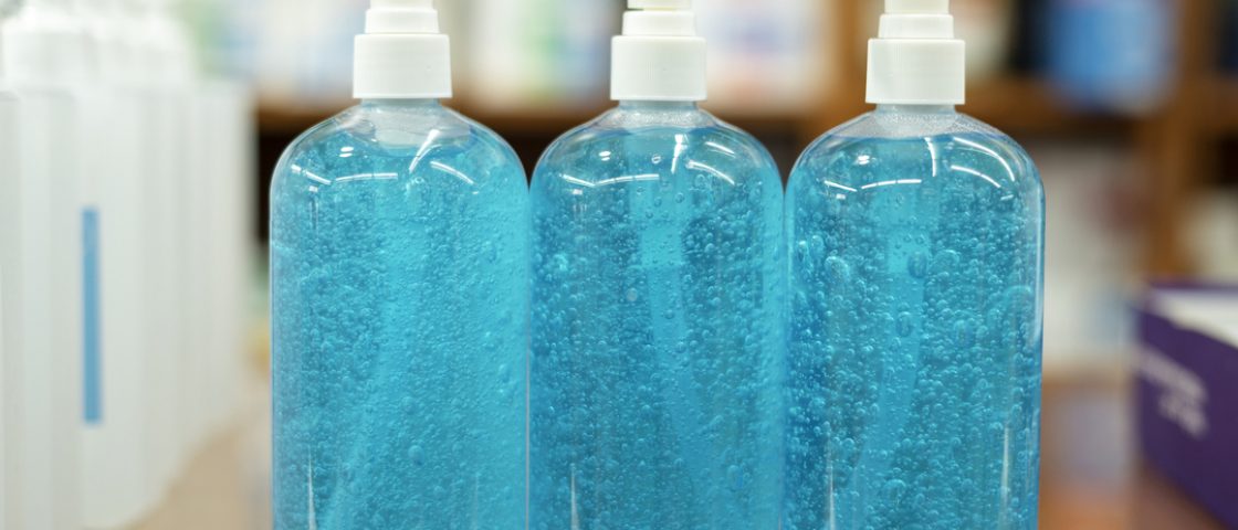 Foto de três frascos de álcool gel em cima de balcão. Ambos são iguais, com pote transparente e conteúdo azul.