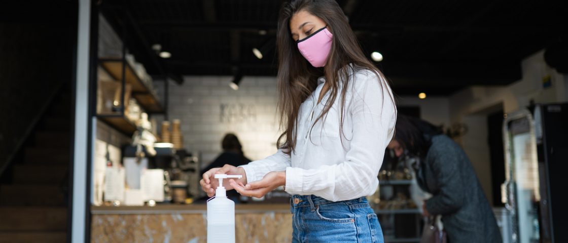 Mulher em um comércio com máscara sobre nariz e boca. Ela aperta a válvula de um recipiente de álcool gel.